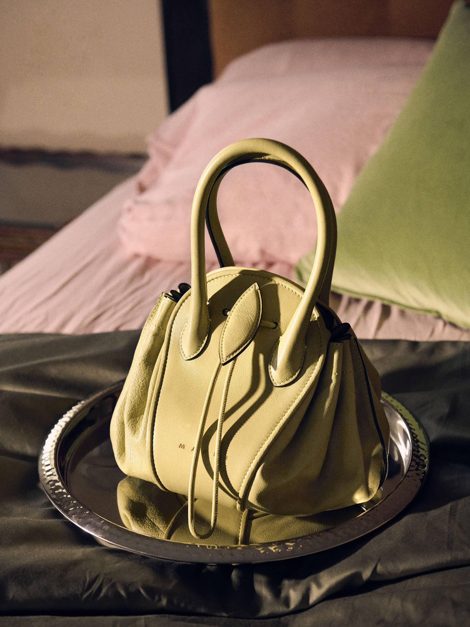 Luxurious yellow handbag on a silver tray with pink and green pillows in the background.